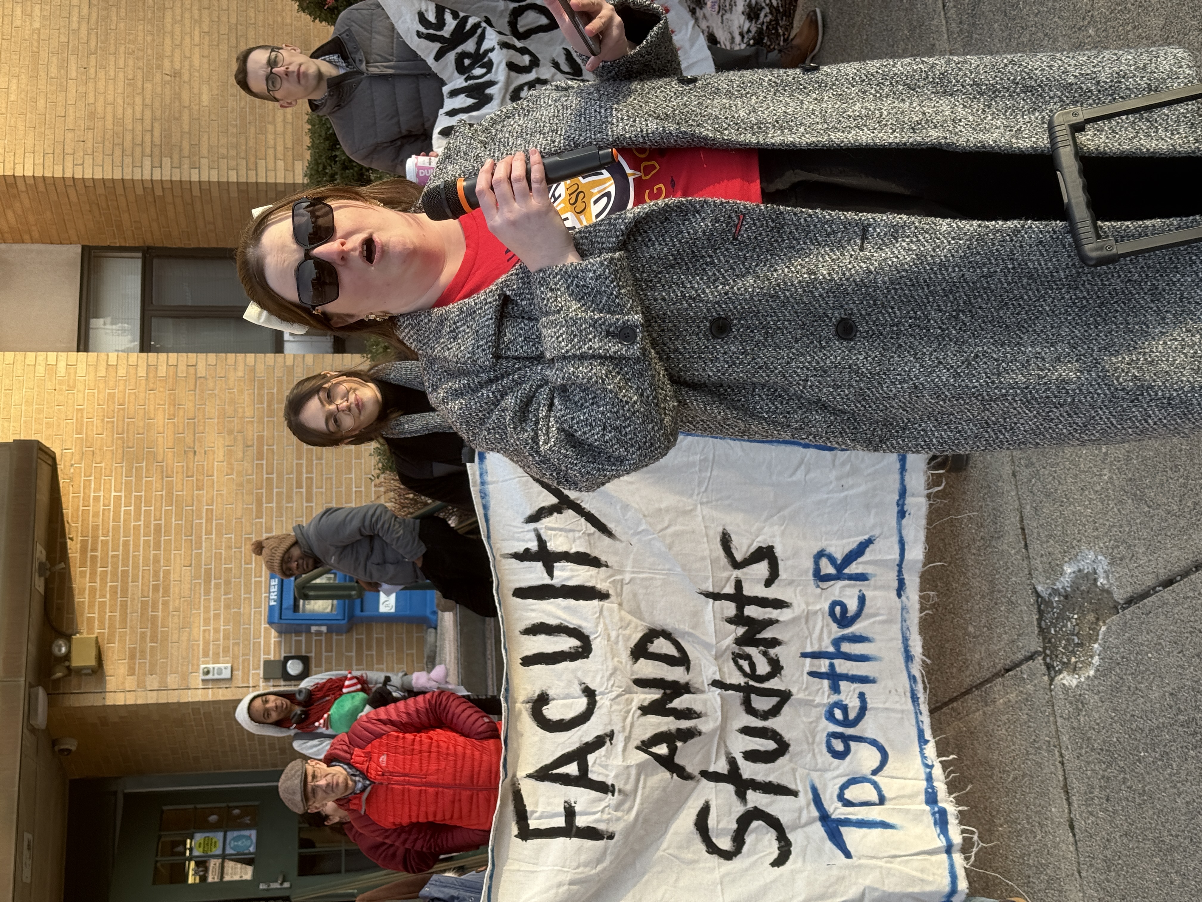 Prof. Ashley Green reads some of the comments from people who signed the petition. Prof. Ashley Green stands in front of a crowd holding signs with a microphone.